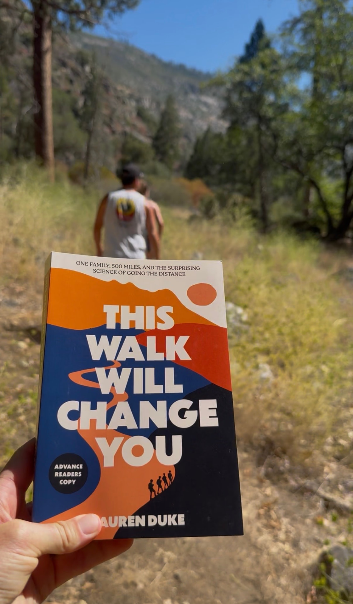 The book held on a wooded trail as a walker heads up the path